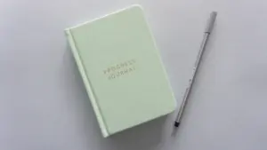 Jasnozielony notes z napisem Progress Journal leżący obok długopisu na szarym tle.